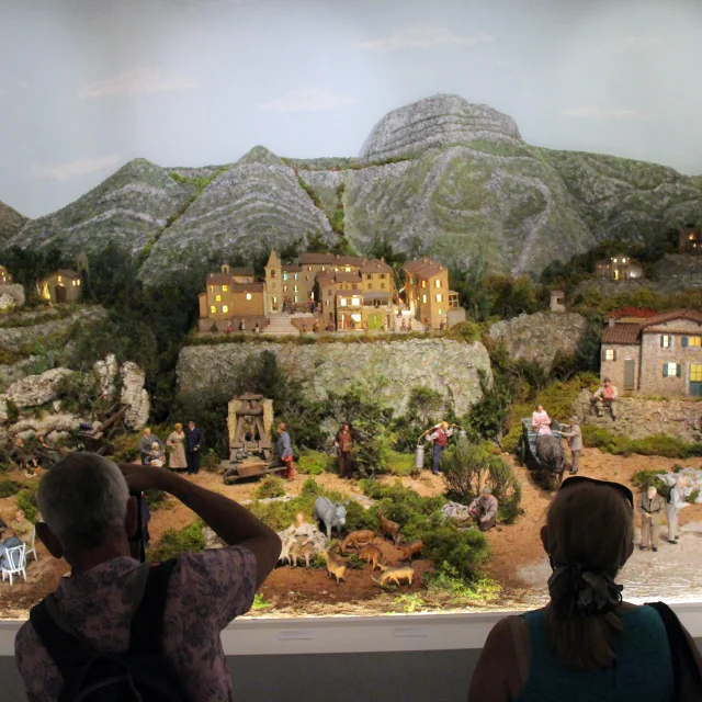 Diorama de village provençal avec maisons miniatures, collines rocheuses et personnages, observé par des visiteurs en intérieur au Petit Monde de Marcel Pagnol à Aubagne