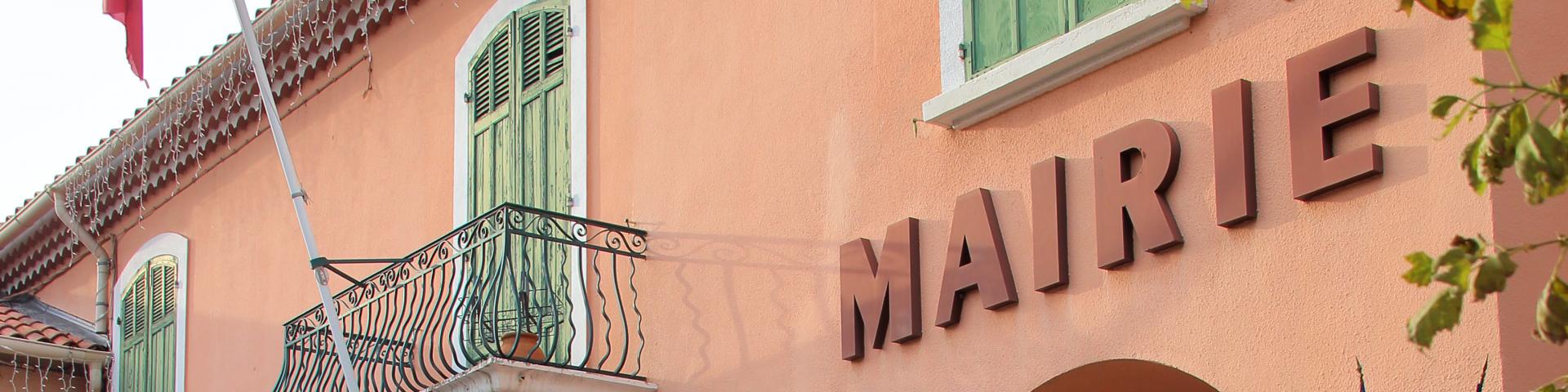 Façade de la mairie de la Destrousse aux murs roses avec volets verts, balcon en fer forgé et drapeaux français accrochés à l’entrée.