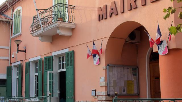 Façade de la mairie de la Destrousse aux murs roses avec volets verts, balcon en fer forgé et drapeaux français accrochés à l’entrée.