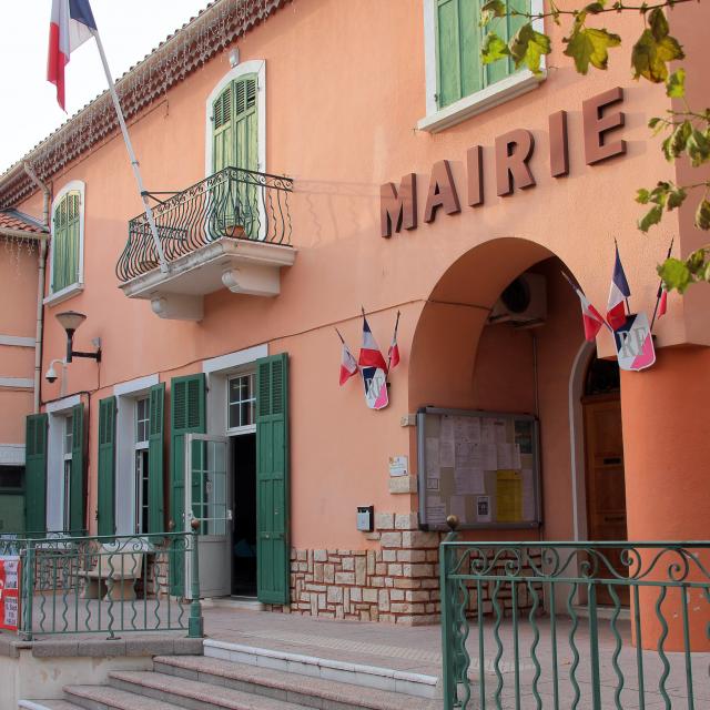 Façade de la mairie de la Destrousse aux murs roses avec volets verts, balcon en fer forgé et drapeaux français accrochés à l’entrée.