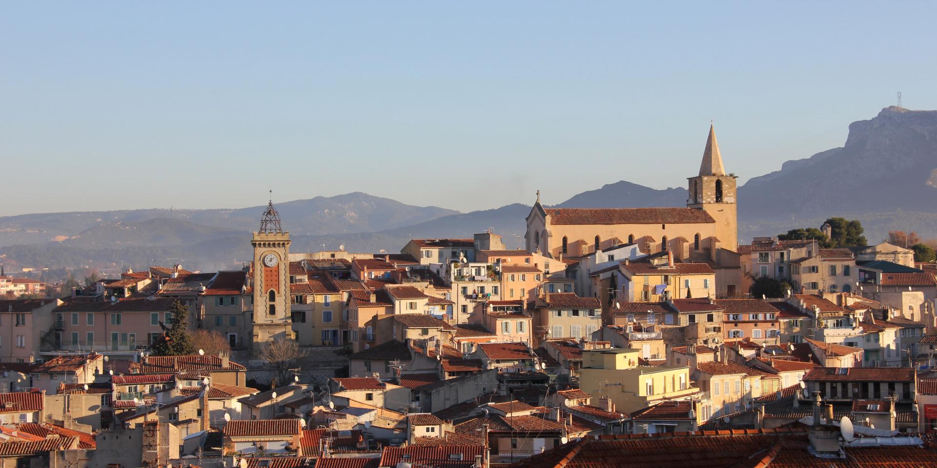 Vue panoramique d’Aubagne montrant les toits de la ville, l’église et la tour de l’Horloge, avec les montagnes en arrière-plan.