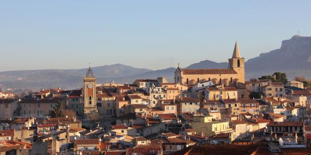Vue panoramique d’Aubagne montrant les toits de la ville, l’église et la tour de l’Horloge, avec les montagnes en arrière-plan.