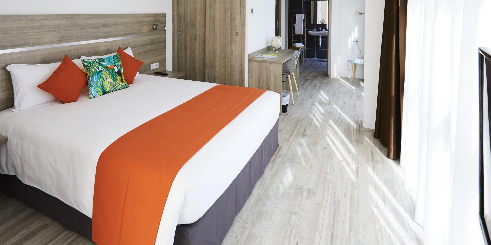 Chambre moderne avec lit double, linge de lit blanc et orange, coussins décoratifs, sol clair et coin cuisine visible en arrière-plan à l'appart hôtel la Girafe à Aubagne
