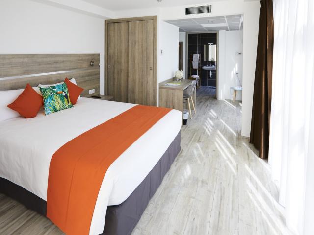 Chambre moderne avec lit double, linge de lit blanc et orange, coussins décoratifs, sol clair et coin cuisine visible en arrière-plan à l'appart hôtel la Girafe à Aubagne