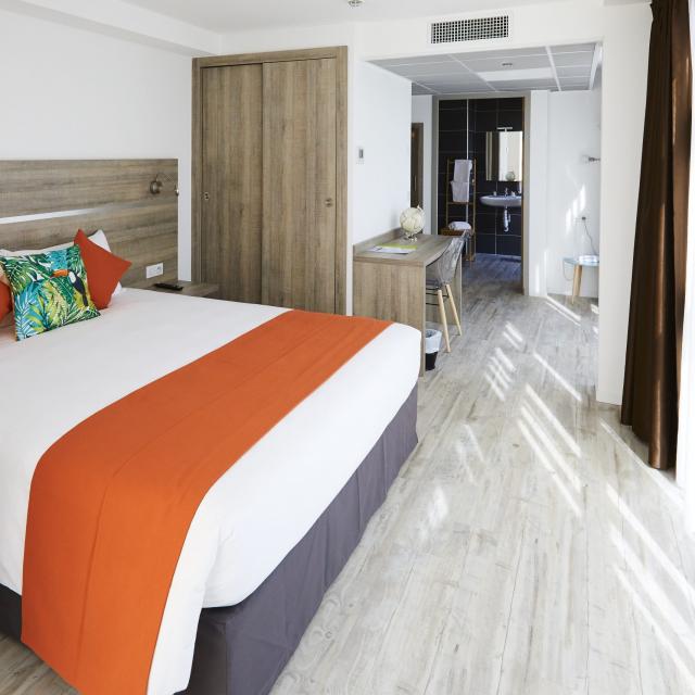 Chambre moderne avec lit double, linge de lit blanc et orange, coussins décoratifs, sol clair et coin cuisine visible en arrière-plan à l'appart hôtel la Girafe à Aubagne