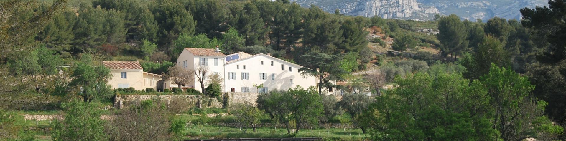 Bastide provençale entourée de prairies et de végétation méditerranéenne, avec une passerelle en bois et des collines boisées surmontées de reliefs rocheux sous un ciel clair au domaine de Font de Mai à Aubagne