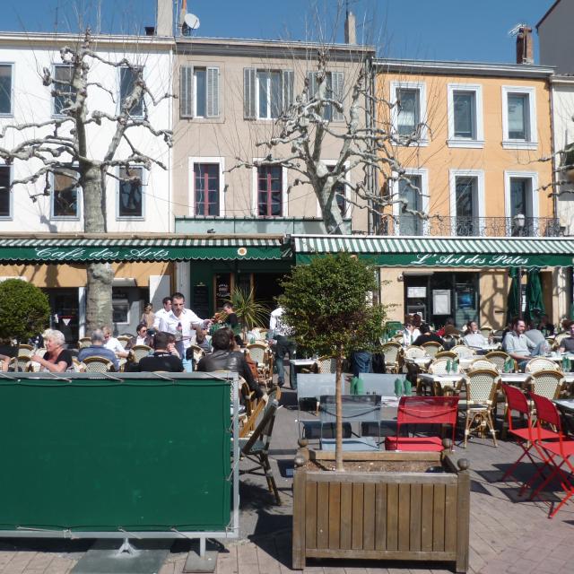 Terrasse de restaurant avec tables et chaises colorées sur une place de village au Café des Arts à Aubagne, clients attablés, façades d’immeubles et arbres taillés en arrière-plan. Café