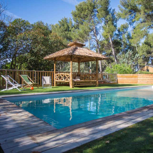 Piscine extérieure avec eau turquoise, terrasse en bois, pergola et transats, entourée de pins et de végétation naturelle au camping du Garlaban à Aubagne