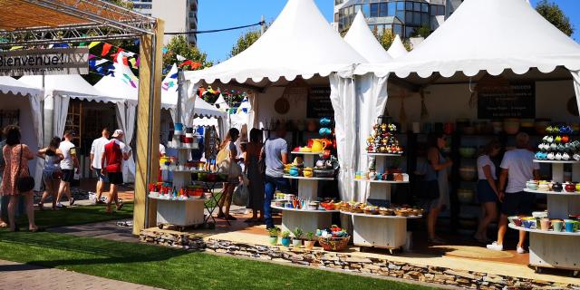 Stands de marché en plein air présentant des poteries et objets en céramique colorés, avec des visiteurs circulant entre les étals sous des tentes blanches.