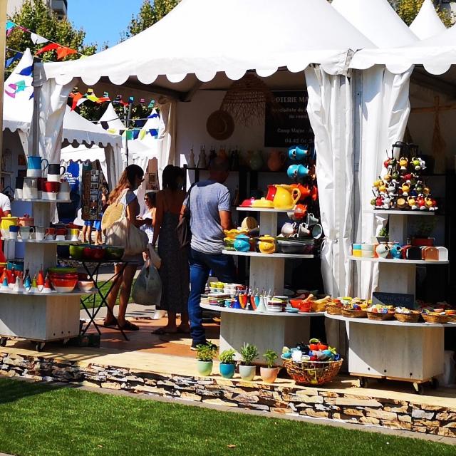 Stands de marché en plein air présentant des poteries et objets en céramique colorés, avec des visiteurs circulant entre les étals sous des tentes blanches.