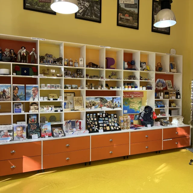 Boutique intérieure avec étagères murales remplies de souvenirs, livres, cartes postales et objets décoratifs, présentés dans un espace lumineux à l'Office de Tourisme d'Aubagne