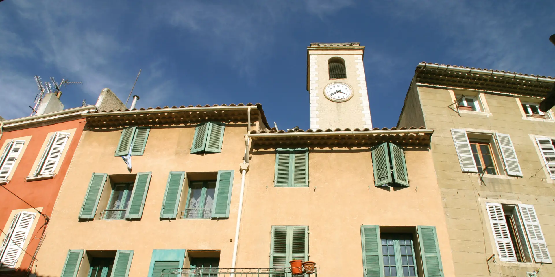 Maisons aux façades ocre avec volets verts et clocher d’église dans un village provençal sous un ciel bleu à Cuges-les-Pins