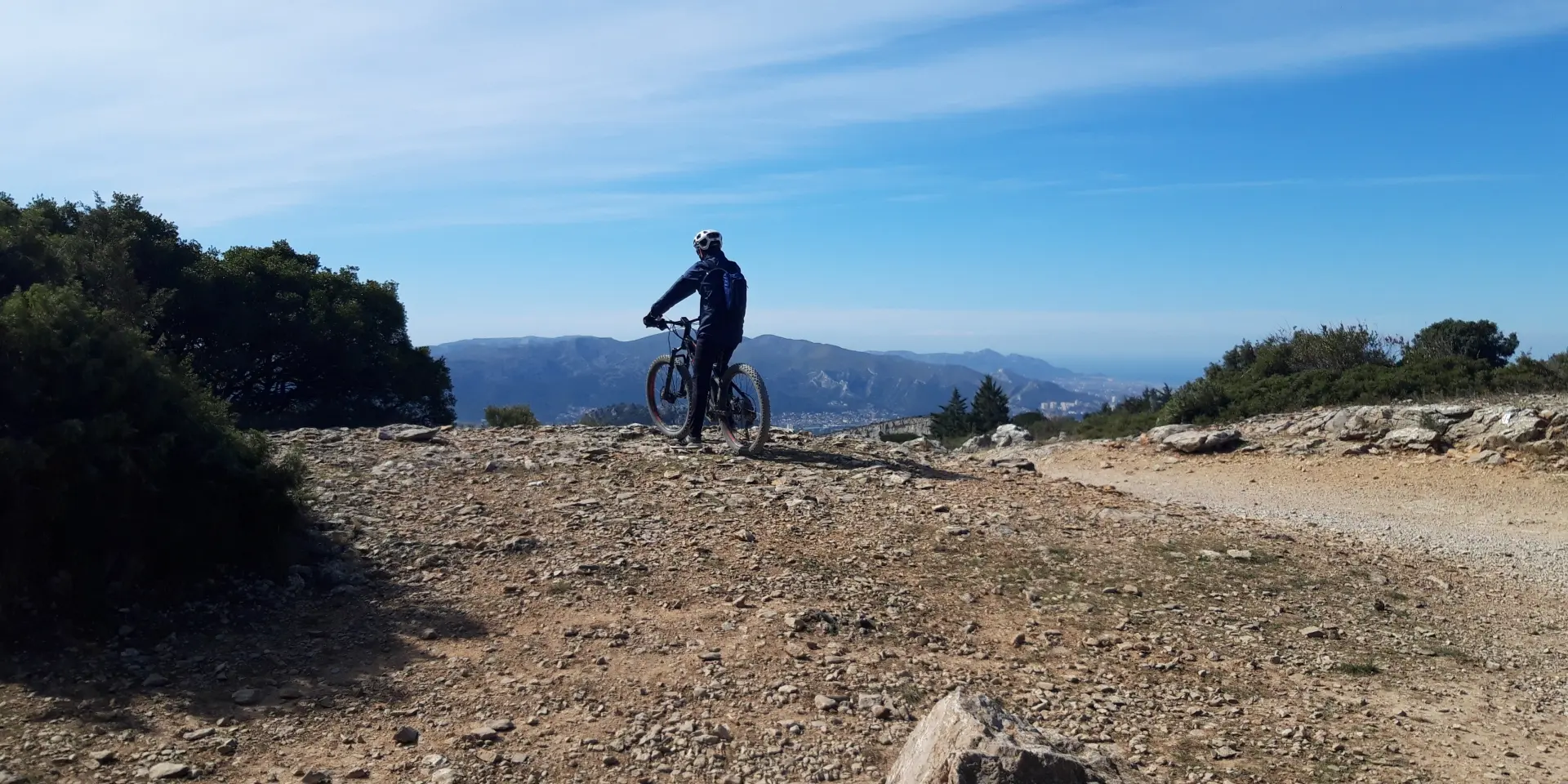 Cycliste en VTT à l'arrêt sur un sentier rocailleux, observant le panorama vers Aubagne et les montagnes aux alentours sous un ciel bleu.